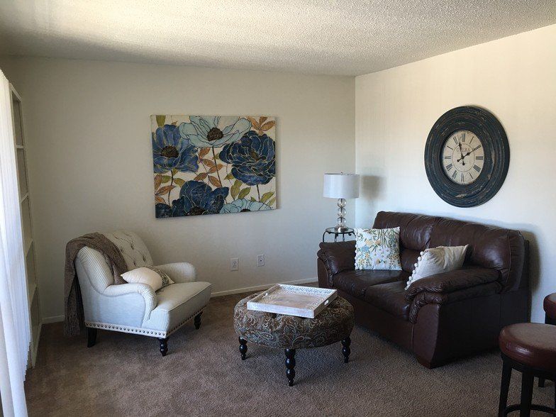 Living room with brown leather sofa, white armchair, blue floral artwork, and a large wall clock.