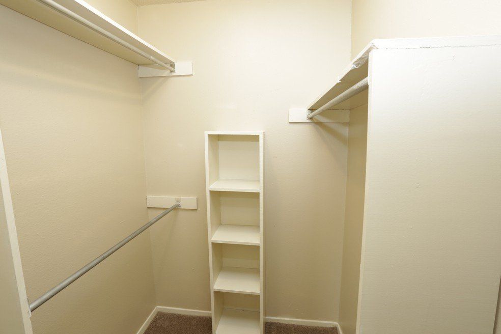 Empty walk-in closet with shelves and rods, beige walls, and brown carpet.