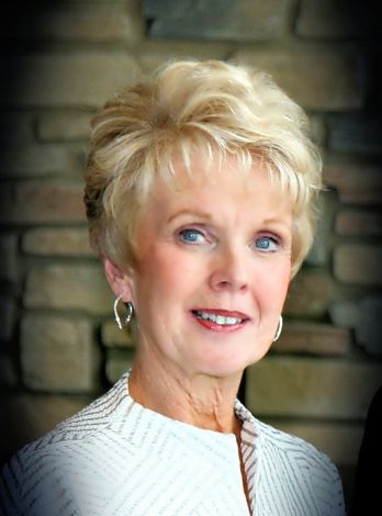 Portrait of a smiling woman with short blonde hair in a white jacket against a stone wall background