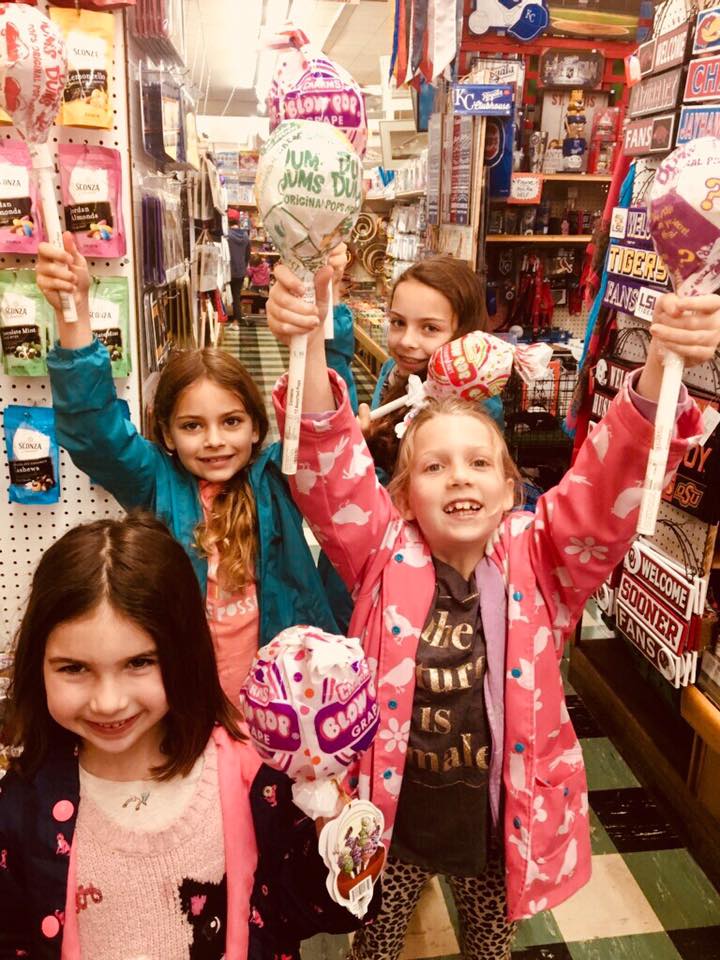 Four children smile while holding large lollipops in a store aisle filled with candy and retail products.