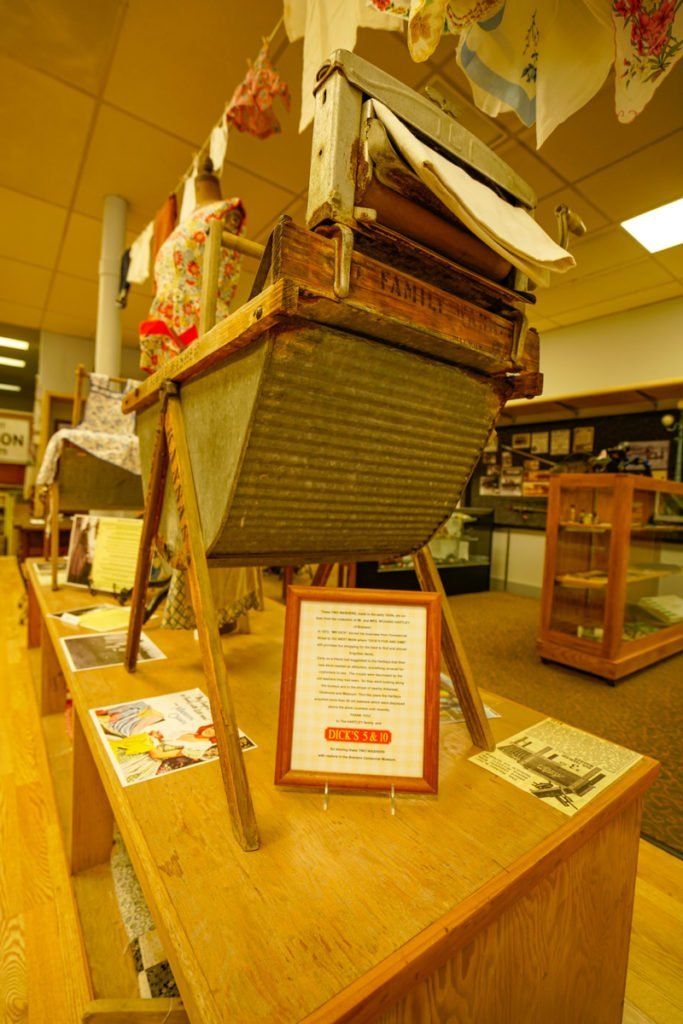 An antique metal washboard basin on a wooden frame stands on a display table inside a museum gallery.