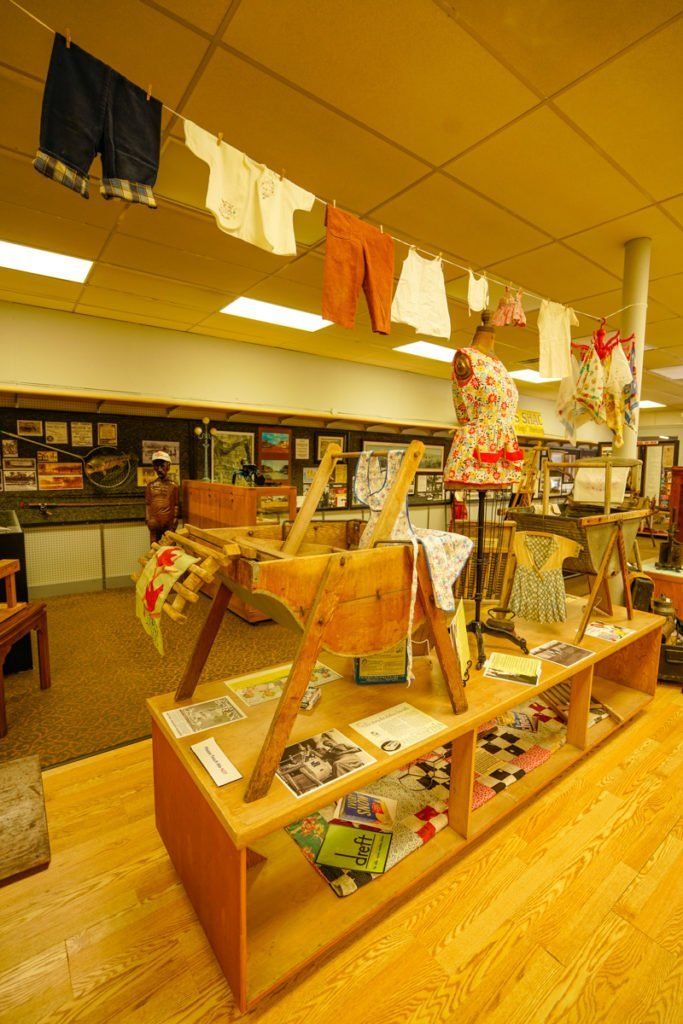 Baby clothes hang from a line above a display table featuring a wooden high chair and mannequin in a brightly lit store.