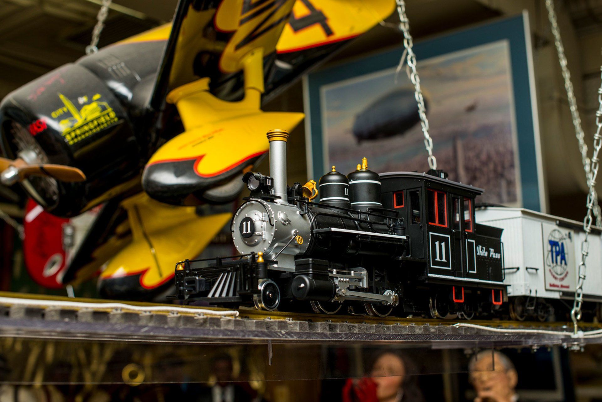 A model steam locomotive sits on a shelf suspended by chains next to a model airplane and a framed airship print.