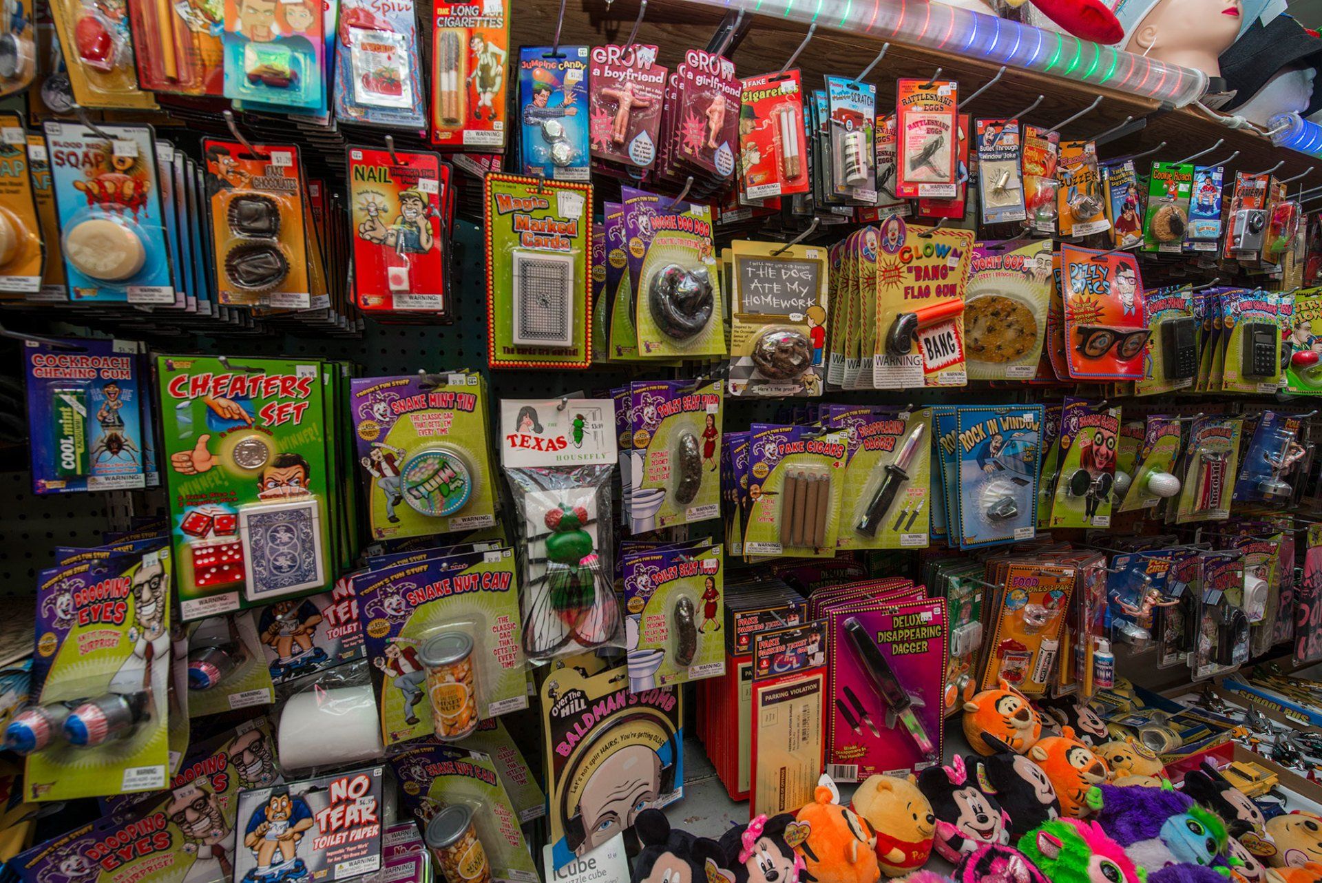 A wall display in a store stocked with various small novelty toys and packaged prank items in colorful hanging boxes.