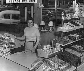 A person behind a checkout counter in a store, with shelves of candy and items visible through a large window.