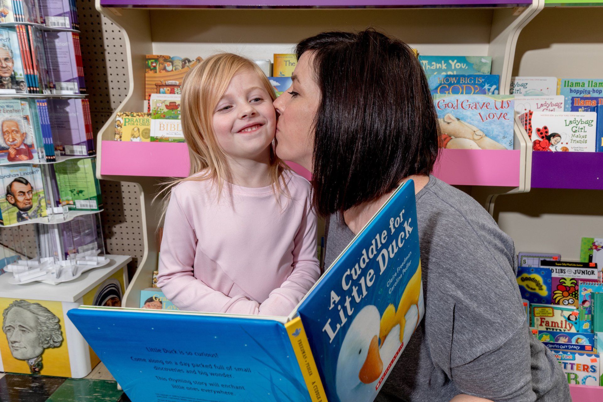 A woman kisses a child on the cheek while holding open a picture book titled 