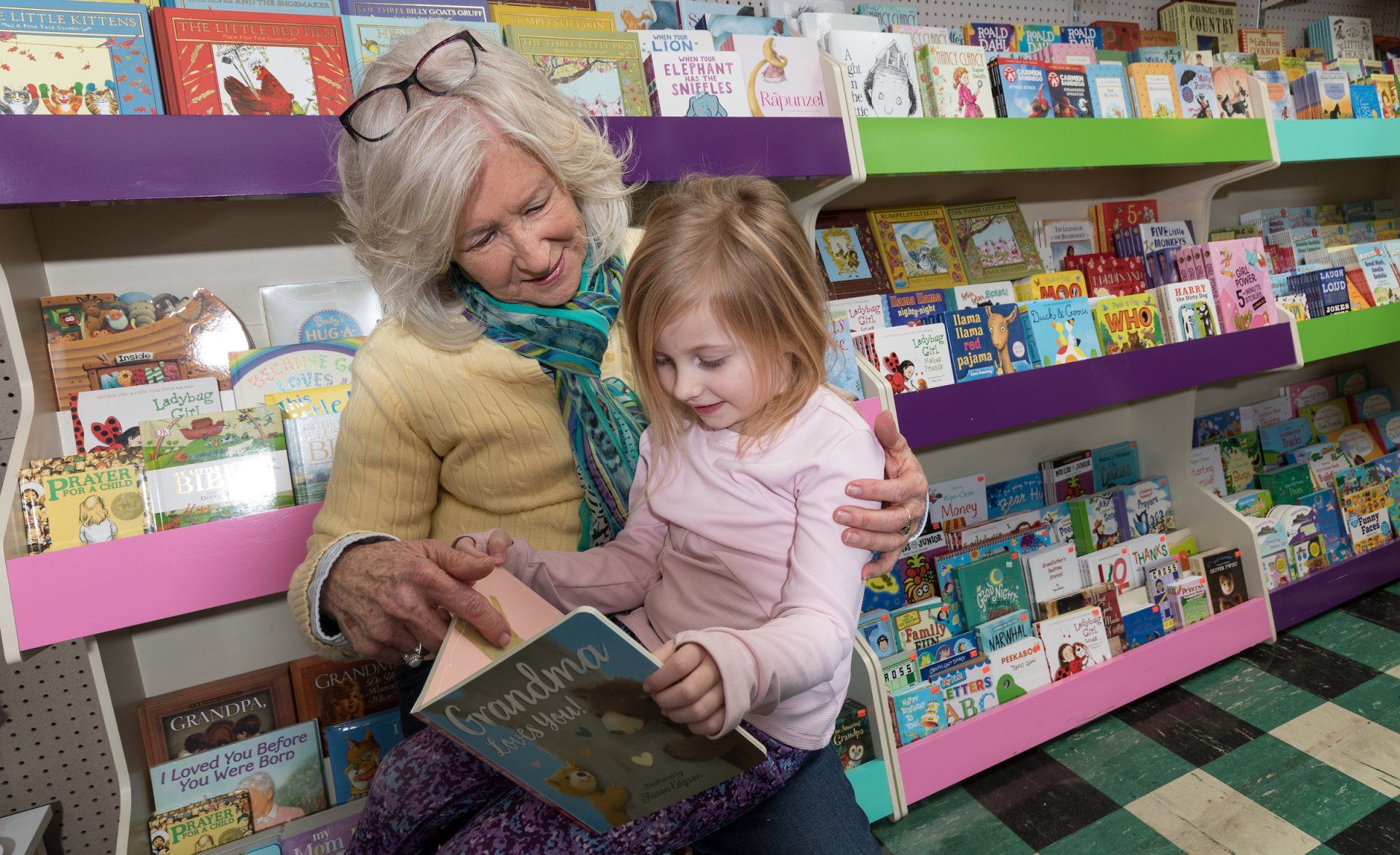 An adult and child sit together in a colorful bookstore, looking at an open book with focused, pleasant expressions.