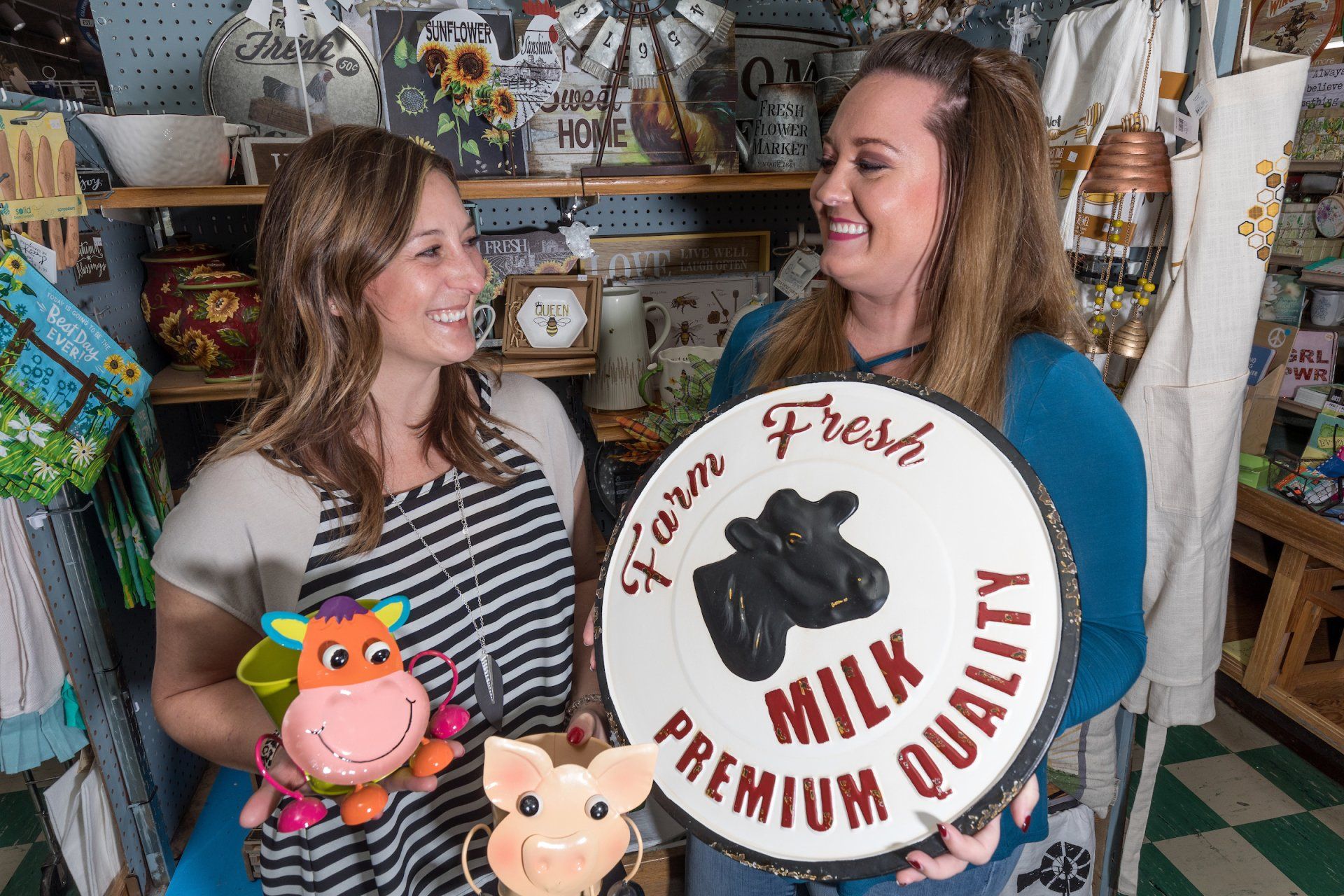 Two people in a retail shop holding farm-themed decor, including a sign that reads 