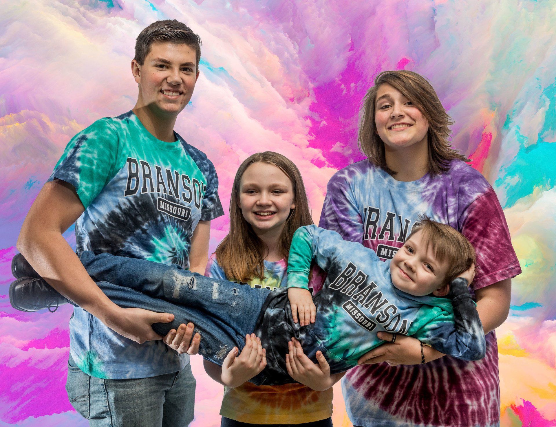 Four people in matching tie-dye shirts pose against a colorful background, with one person held horizontally by two others.
