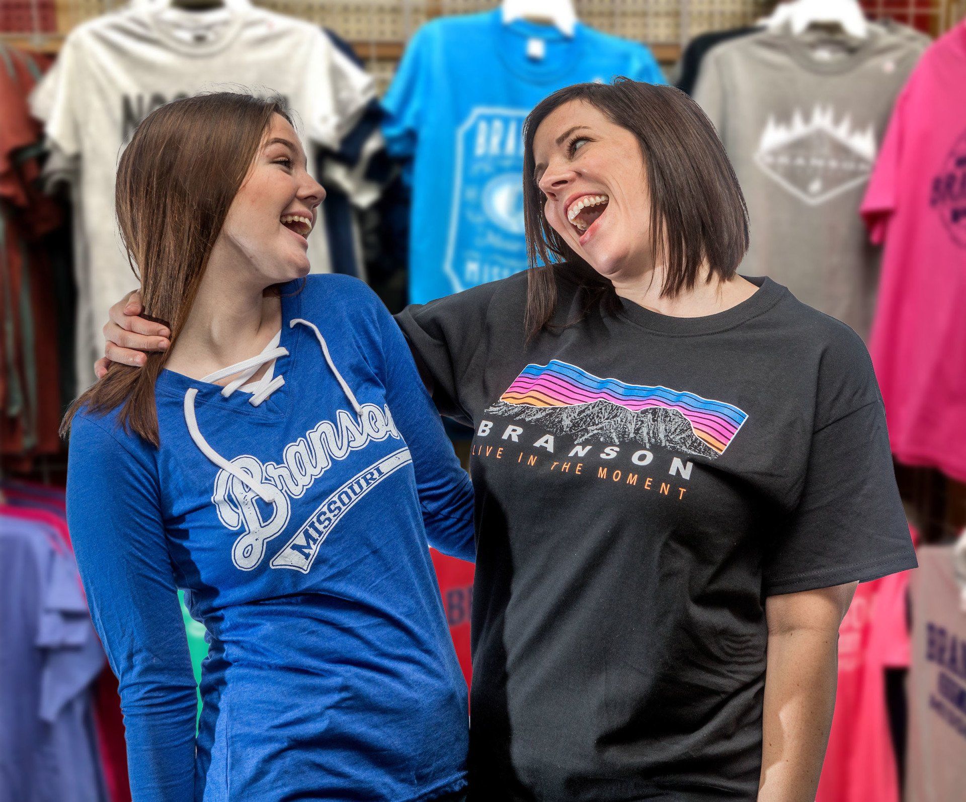 Two smiling people wearing Branson-branded shirts stand in front of a rack of colorful t-shirts in a store.