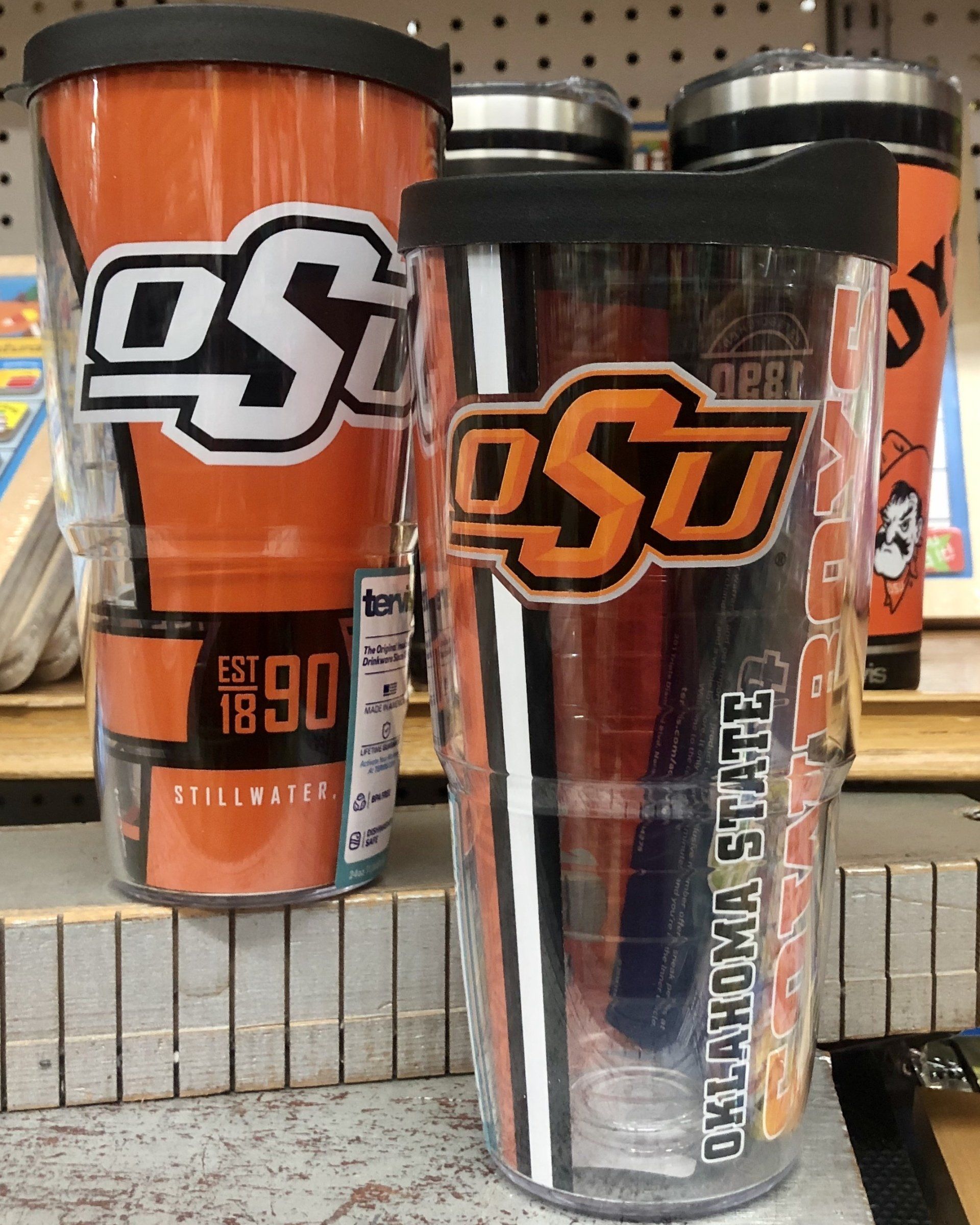Two Oklahoma State University tumblers, one solid orange with a white logo and one clear with an orange logo, on a shelf.