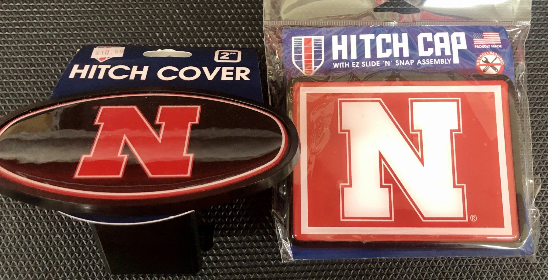 Two University of Nebraska trailer hitch covers, one oval and one rectangular, side by side on a dark surface.