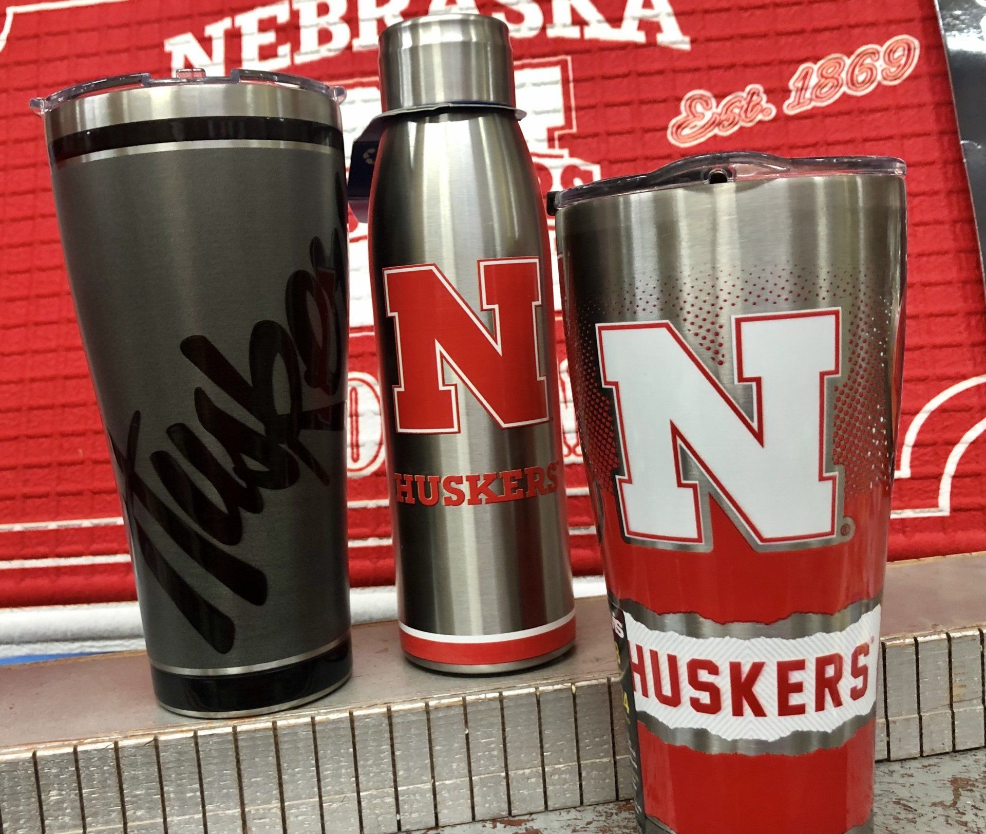 Three University of Nebraska branded drinkware items: a dark grey tumbler, a silver water bottle, and a red tumbler.