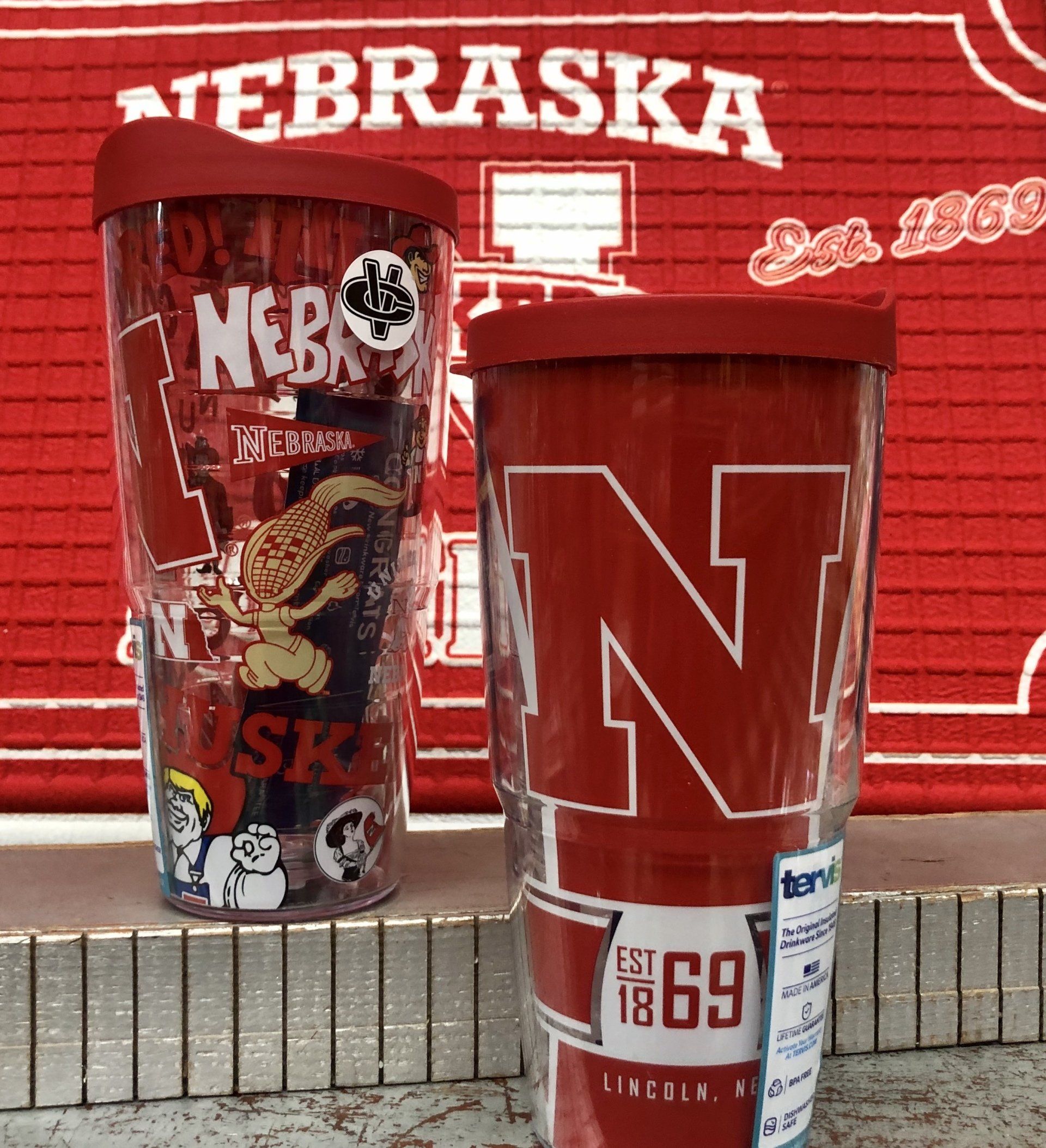 Two University of Nebraska travel tumblers stand in front of a red Nebraska-branded cloth backdrop.