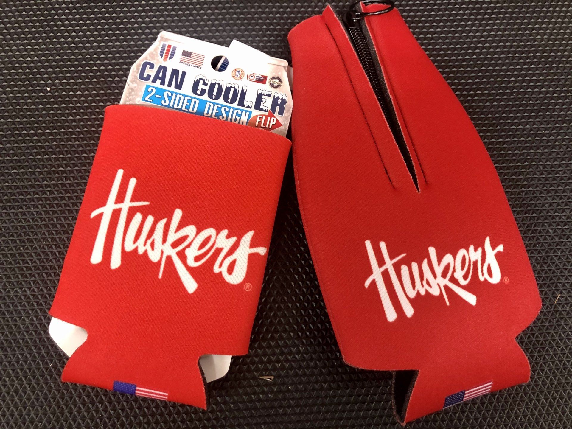 Two red Nebraska Huskers branded foam can coolers, one inside its packaging and one open, on a textured surface.