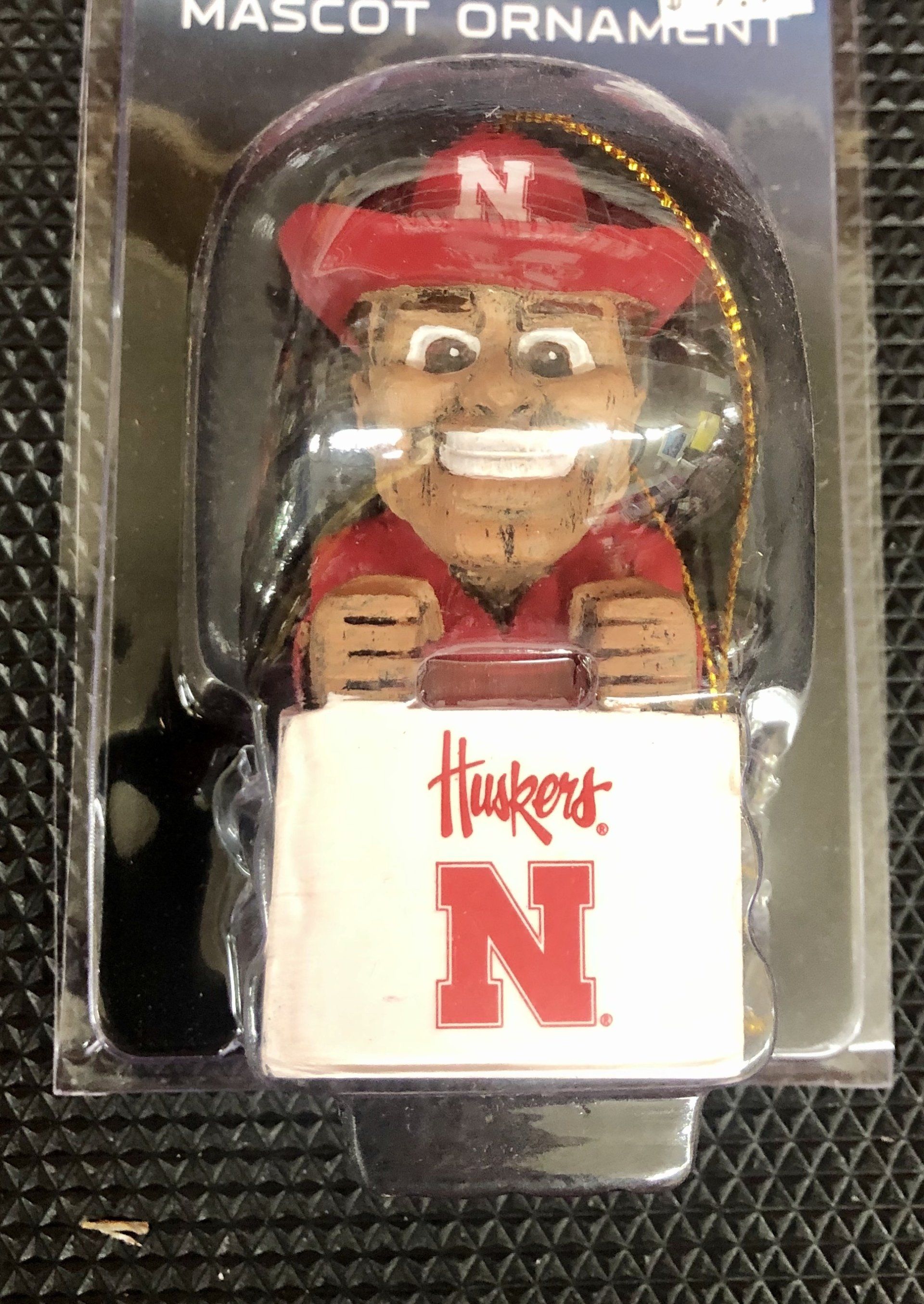 A University of Nebraska Huskers mascot ornament in its plastic packaging, featuring the character in a red hat and shirt.