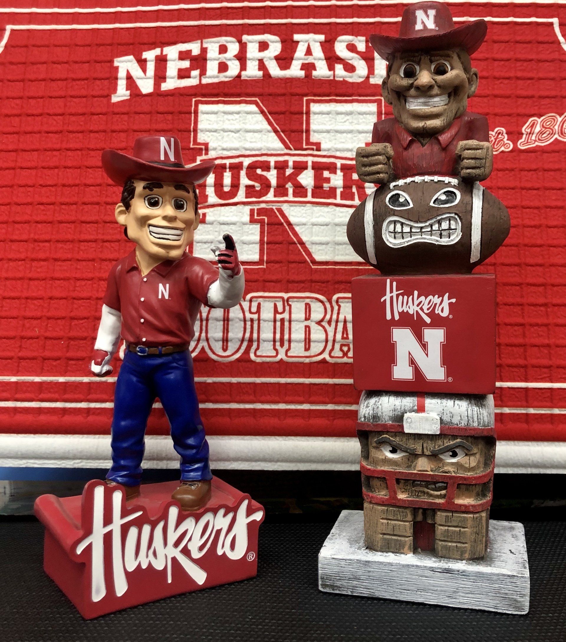 Two Nebraska Cornhuskers bobblehead figurines in front of a team-branded wall hanging.
