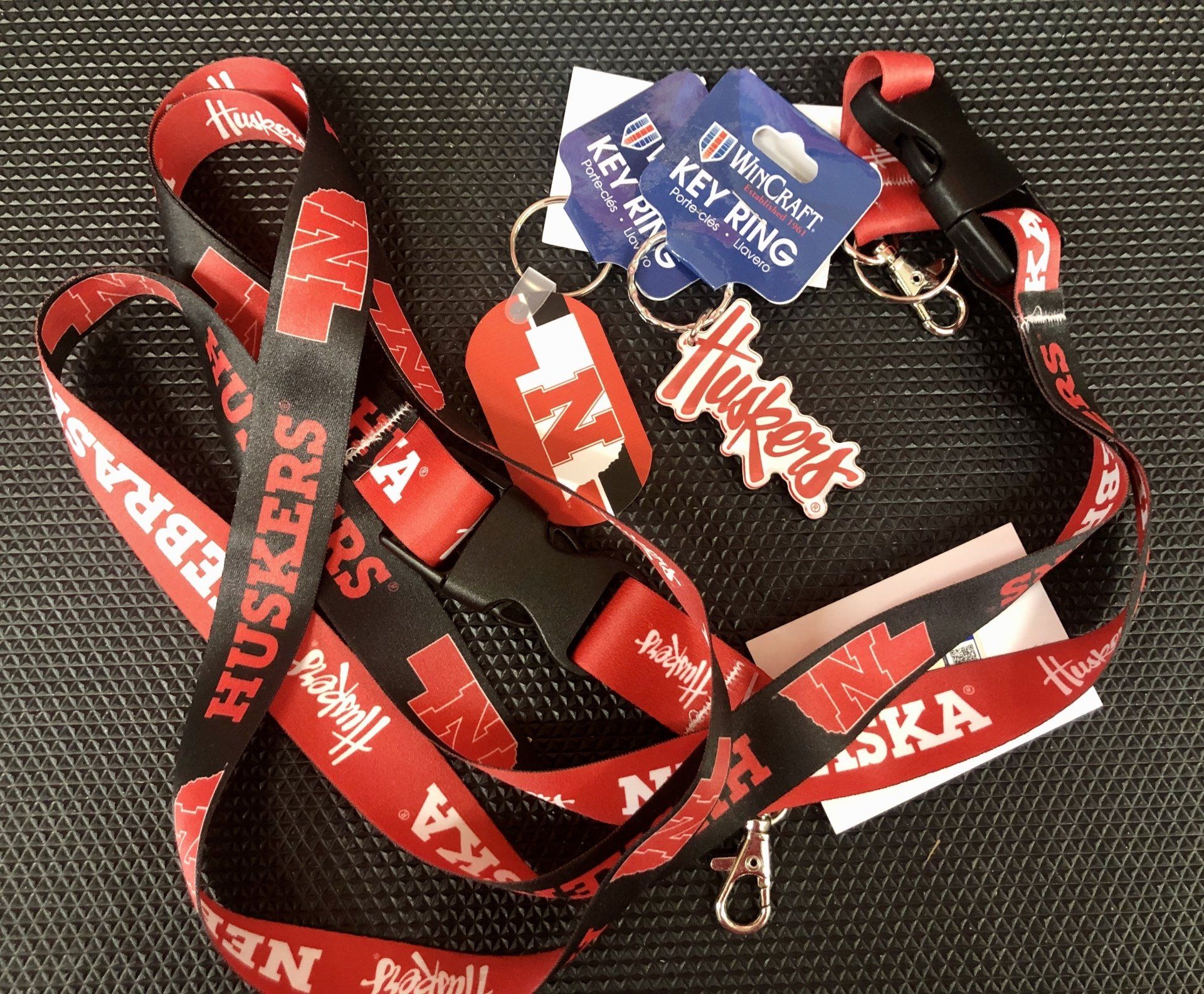 A black and red Nebraska Huskers branded lanyard and two keychains on a textured black surface.