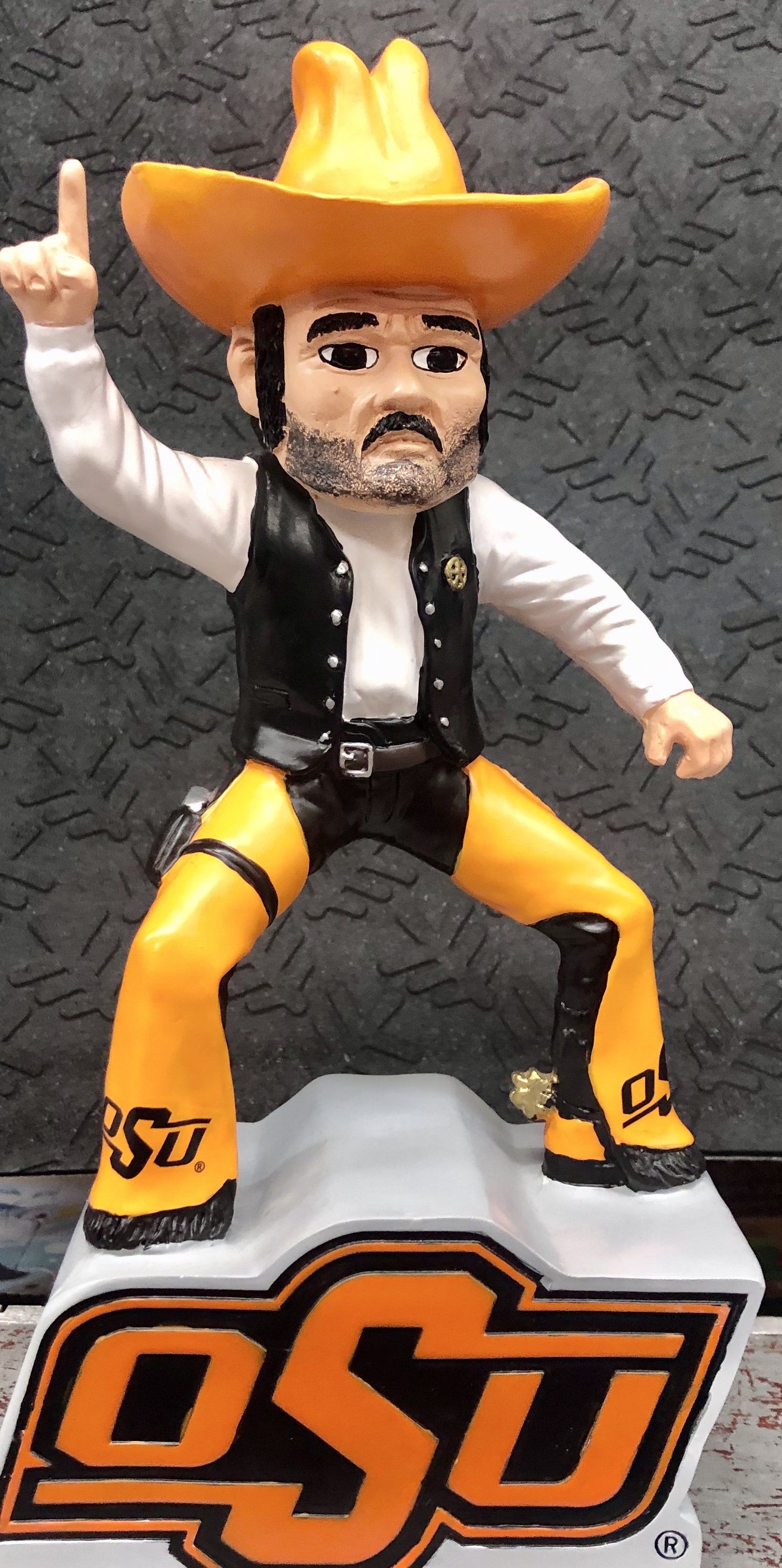 A bobblehead of the Oklahoma State mascot, Pistol Pete, wearing a cowboy hat and orange chaps, standing on an OSU base.