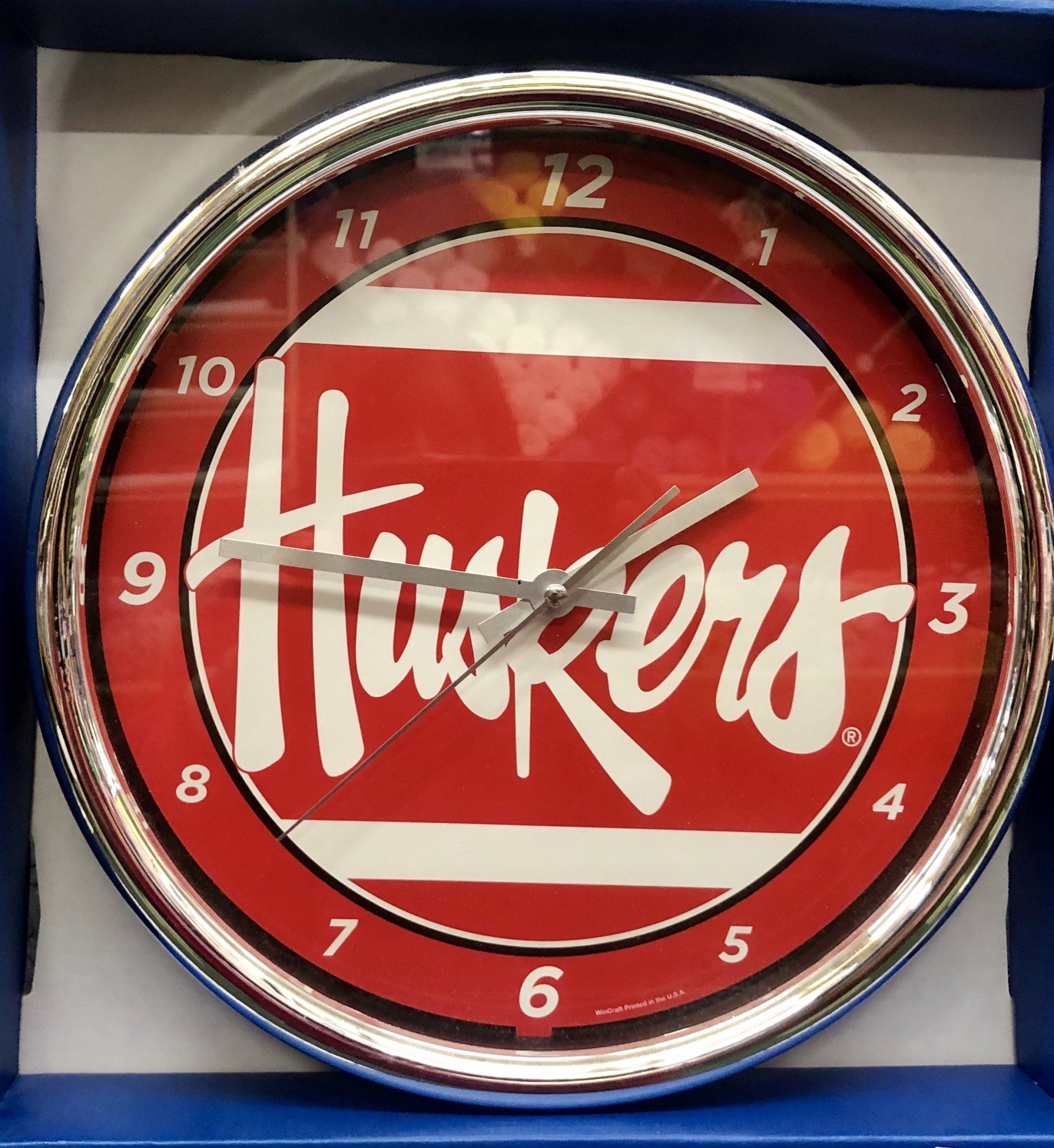 A round, silver-rimmed wall clock featuring a red face with white Nebraska 