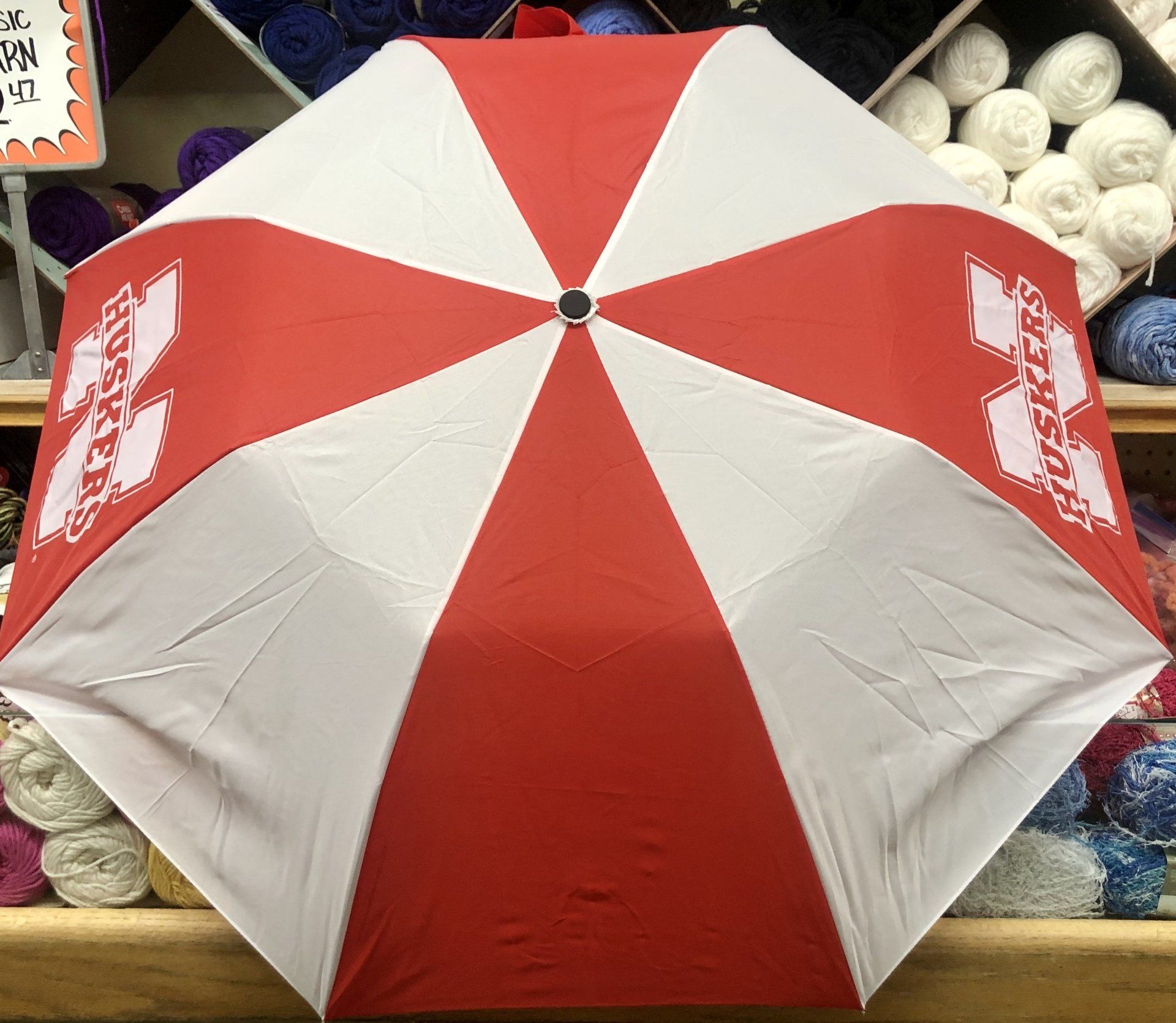 An open red and white striped umbrella featuring the Nebraska Huskers logo, displayed in a store with yarn in the back.