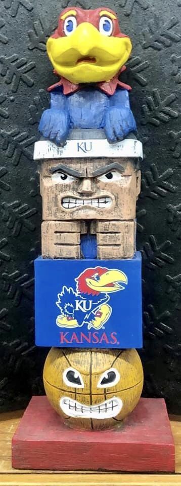 A vertical wooden totem featuring the KU Jayhawk, a fierce face, the Kansas logo, and a basketball, on a red base.