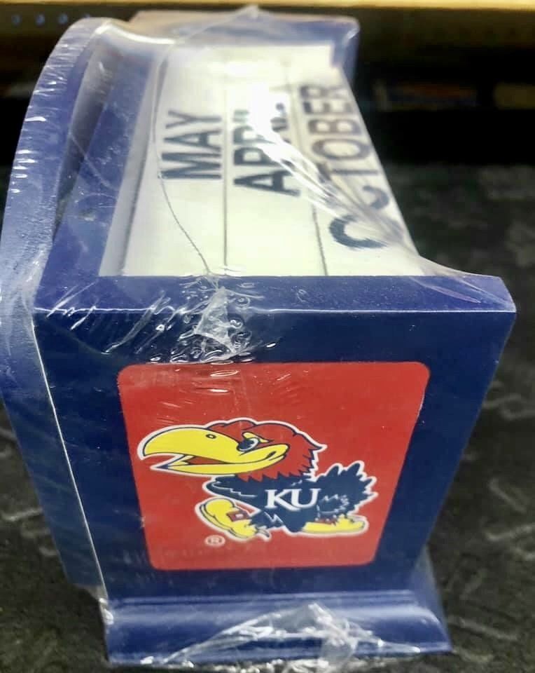 A Kansas Jayhawks desk calendar wrapped in plastic, featuring the blue and red team logo on a blue base.