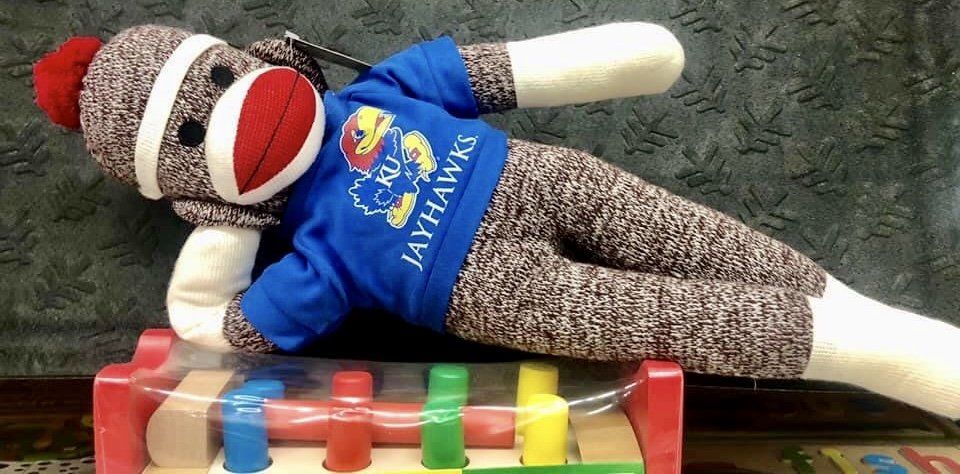 A plush sock monkey in a blue KU Jayhawks t-shirt reclines on top of a colorful wooden toy pounding bench.