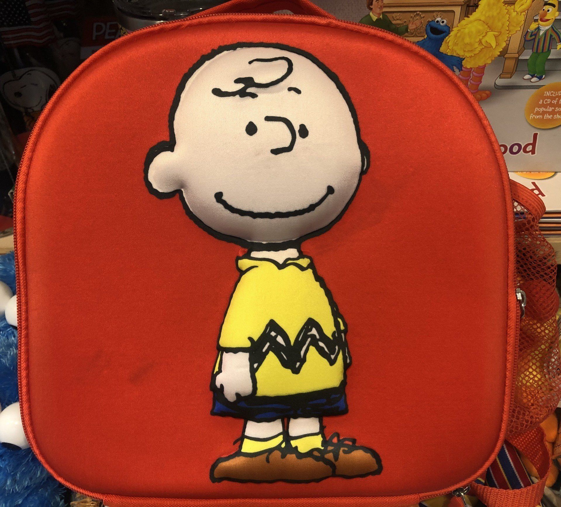 A bright orange lunchbox featuring a smiling Charlie Brown wearing his signature yellow zigzag shirt and blue shorts.