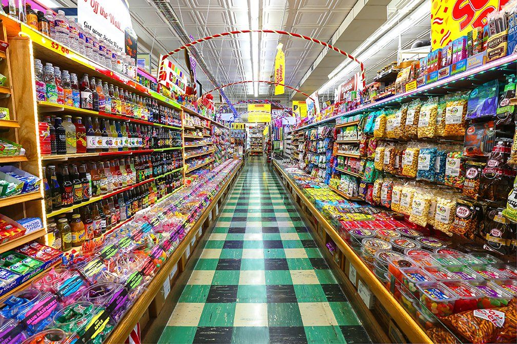 An aisle in a grocery store, lined with shelves of snacks and drinks on both sides and a green-and-white checkered floor.