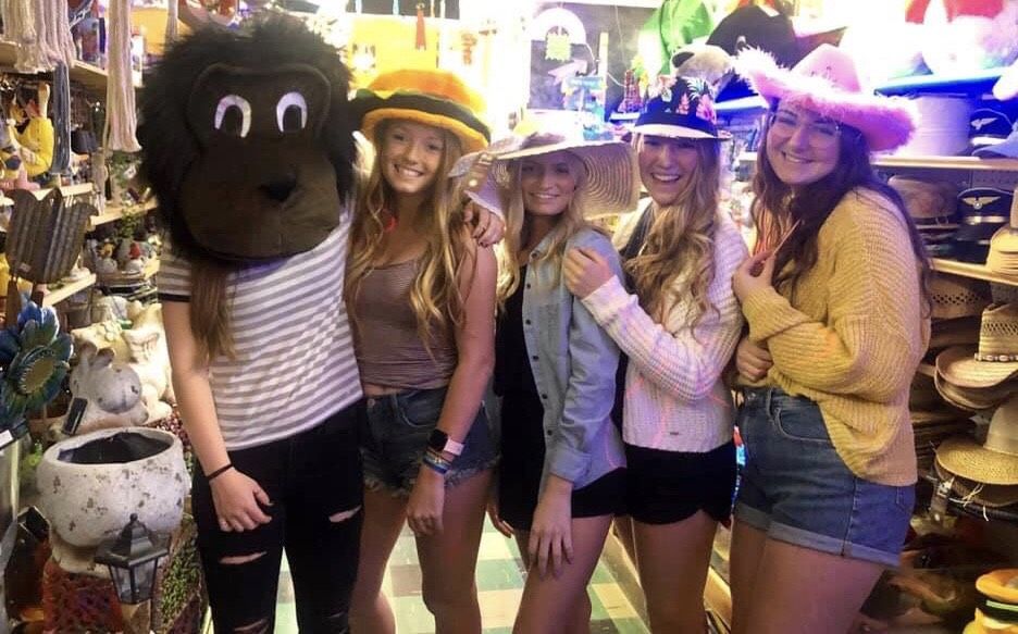 Five people stand in a shop, four smiling while one wears a large gorilla mask; they wear various hats and casual clothes.