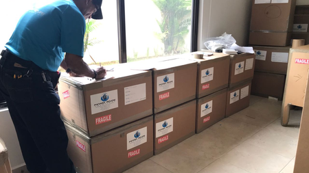 Una persona con camisa azul apila cajas de cartón etiquetadas en una habitación con una ventana grande.