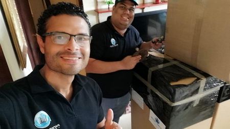 Dos personas sonrientes están de pie junto a un objeto grande embalado en una caja, en una habitación con estanterías y una ventana.