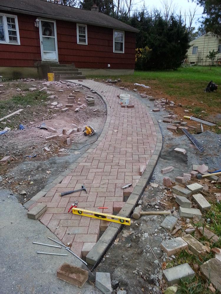 Curved brick walkway under construction beside a red house, with scattered tools and loose pavers.