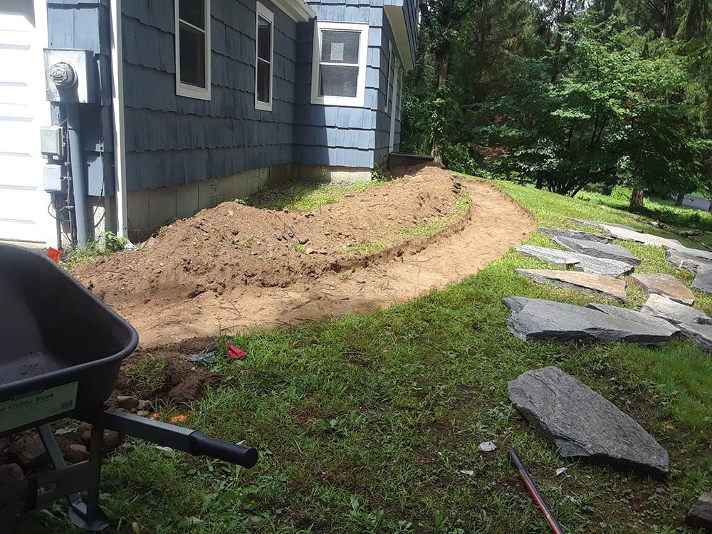 Backyard beside a house with fresh soil, stepping stones, and a wheelbarrow in the grass.