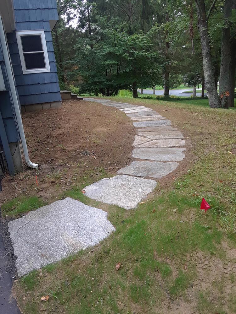 Curved stone stepping path beside a blue house across a grassy yard toward trees.