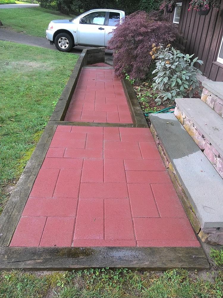 Red brick walkway bordered by wood timbers beside a house, leading to a parked silver car.