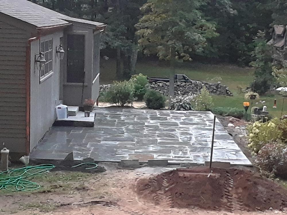 Backyard patio with stone pavers beside a house, garden bed, and soil mound in the foreground