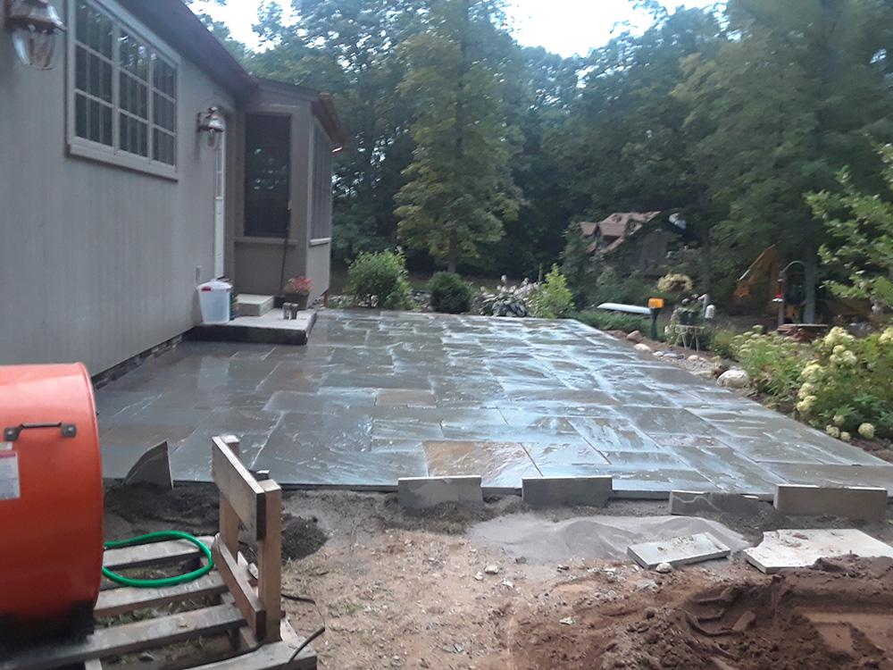 Backyard patio under construction beside a house, with stacked pavers and dirt in the foreground
