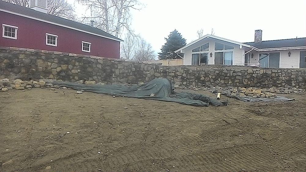 Rural yard with dirt foreground, stone wall, tarped pile, and houses under an overcast sky