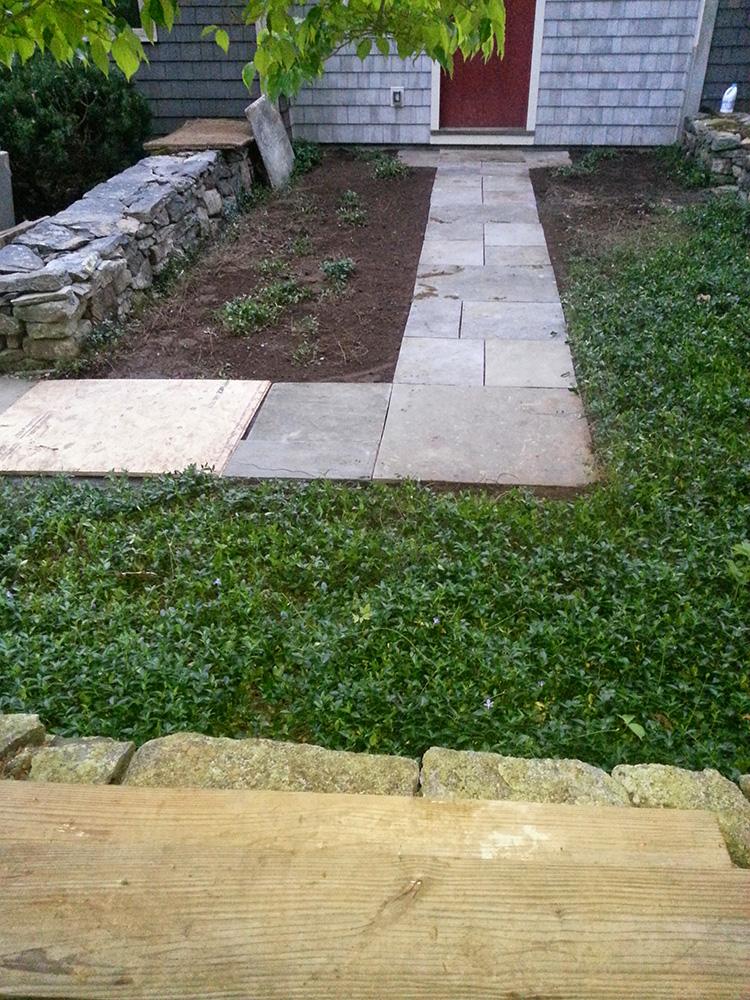 Front yard walkway with stone pavers leading to a house door, bordered by grass and garden beds.