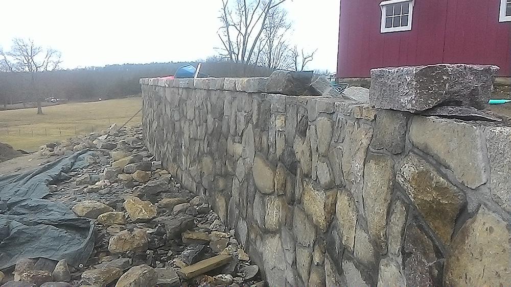 Stone retaining wall beside a red building on a rural hillside, with broken rocks in the foreground