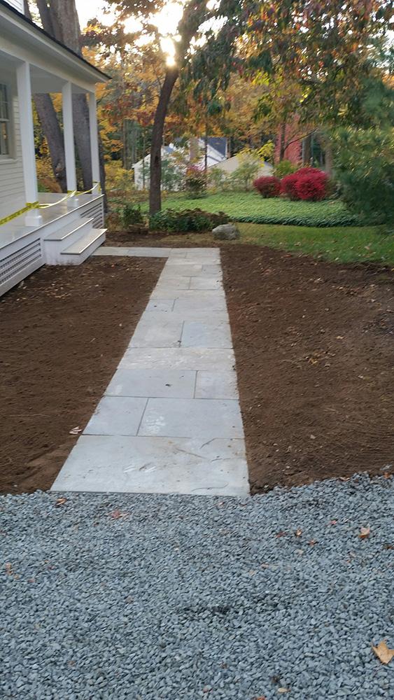 Concrete walkway leading from a house porch to a gravel drive, bordered by bare soil and a garden.
