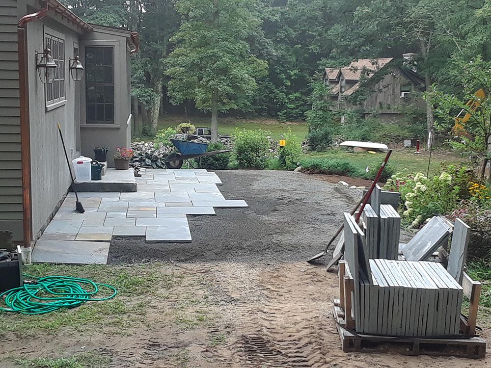 Backyard with paver stones beside a house, tools and stacked materials, and a garden in the background