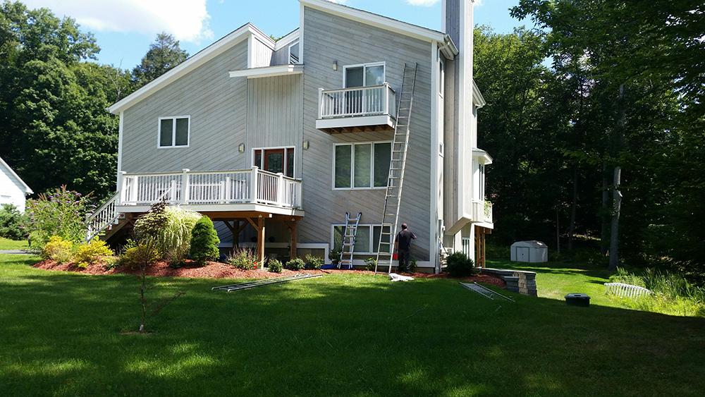 Light gray house with small balconies and a green lawn, backed by trees in bright daylight