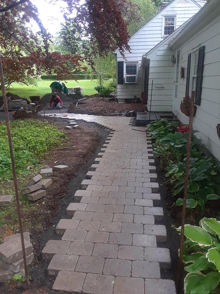 Brick walkway under construction beside a white house, with a worker near the front yard.