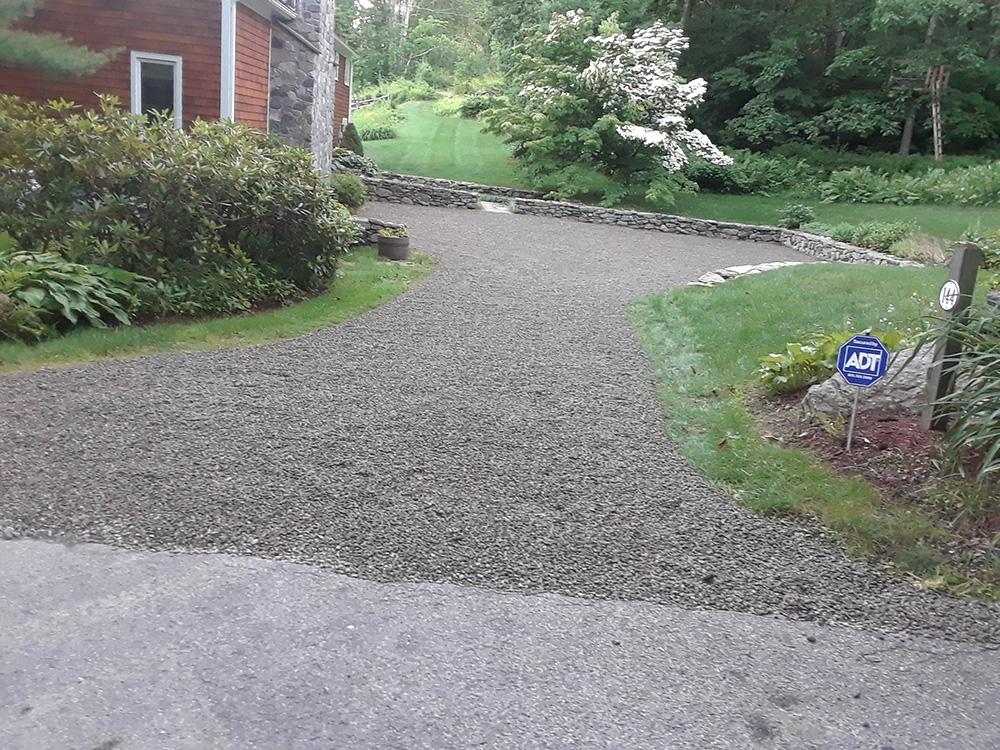 Gravel driveway curving past a brick house into a green garden with shrubs and flowering trees