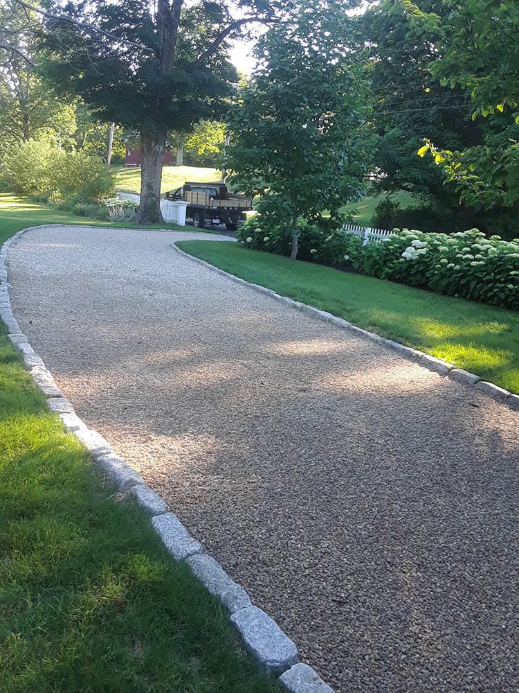 Curved gravel driveway bordered by grass and trees, leading to a parked car and garage in the distance.