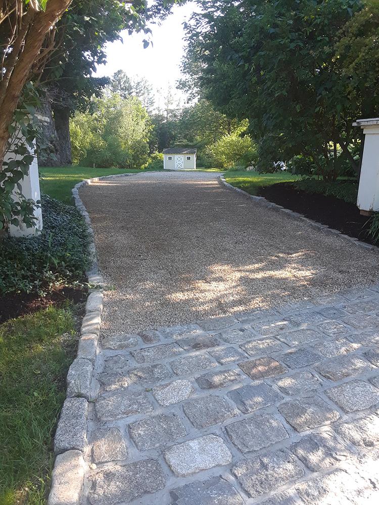 Stone driveway leading through a tree-lined garden toward a white gate or gatehouse.