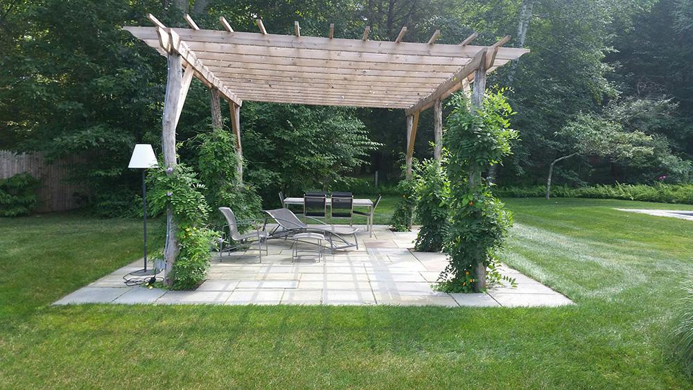 Wooden pergola over a patio seating area surrounded by green lawn and trees.